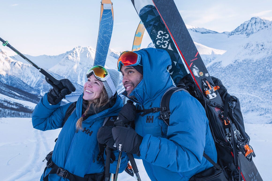 2 people trekking a mountain with skis on their backs and helly hansen gear on, smiling looking up at the mountains