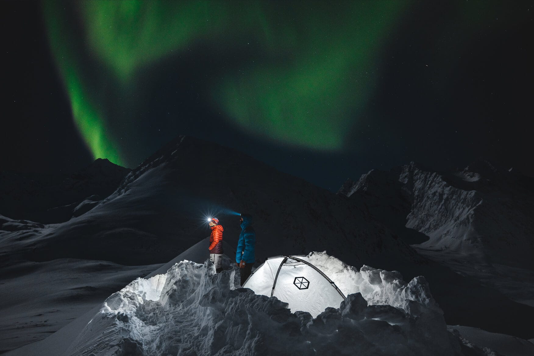2 people with headlamps on standing next to a tent under the northern nights 