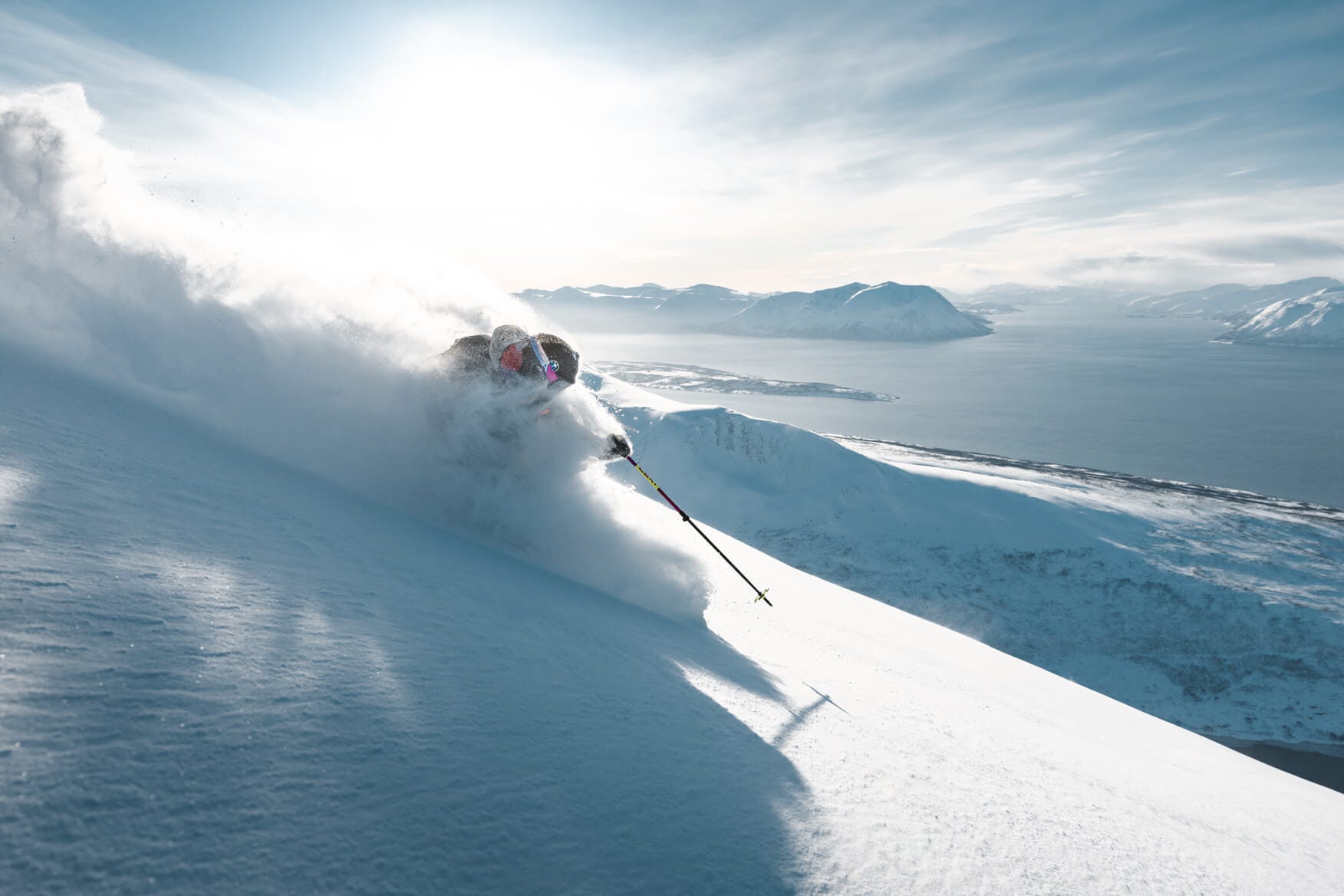 person skiing in the sun in fresh deep powder with a view of the fjord in the background