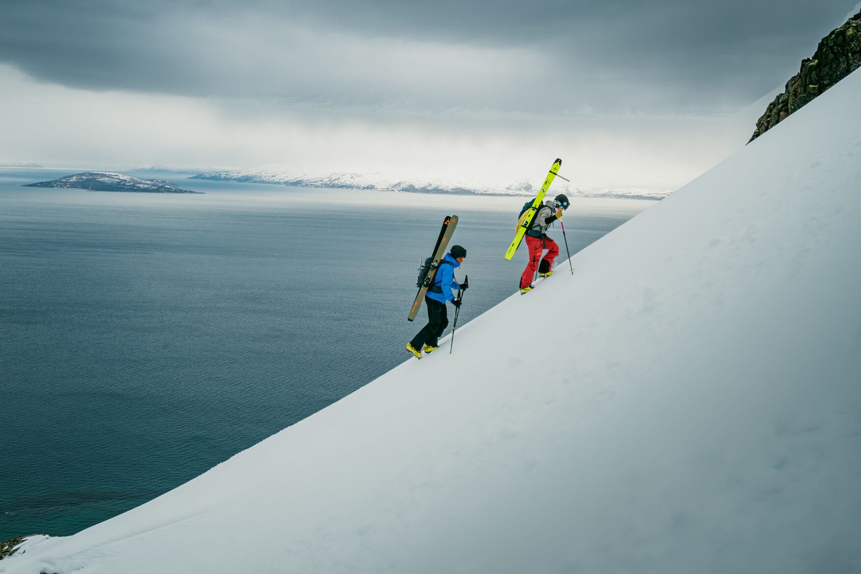 2 people scaling a mountain with skiis on their back in the snow with a view of the fjord in the background
