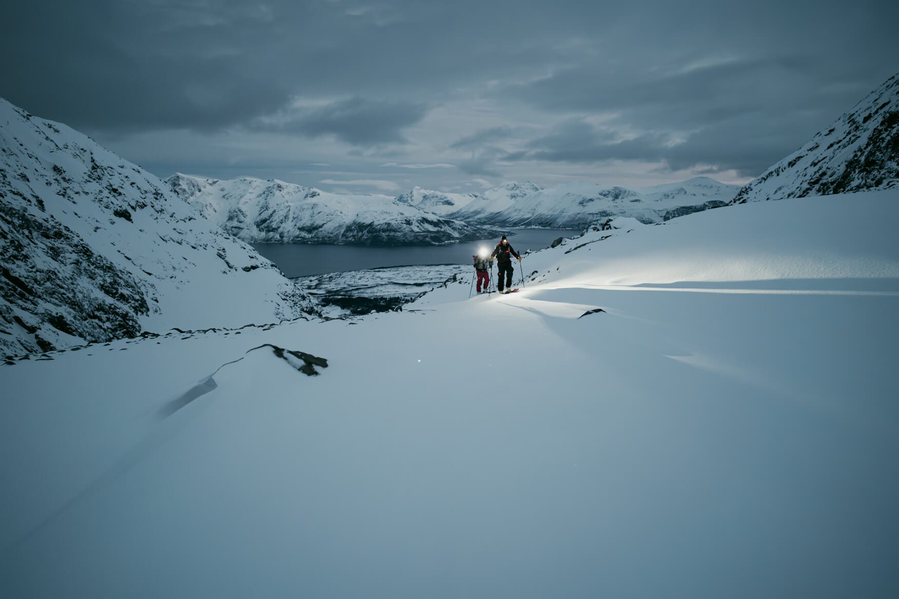 2 people ski touring with a view of a fjord in the evening with headlamps on 